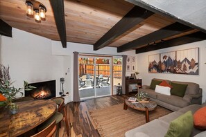 A cozy living room with a fireplace, a sofa, a dining table, and a large window with a view of the outdoors. - Summit Après | 2-Min Walk to Snow Summit (Big Bear Lake)