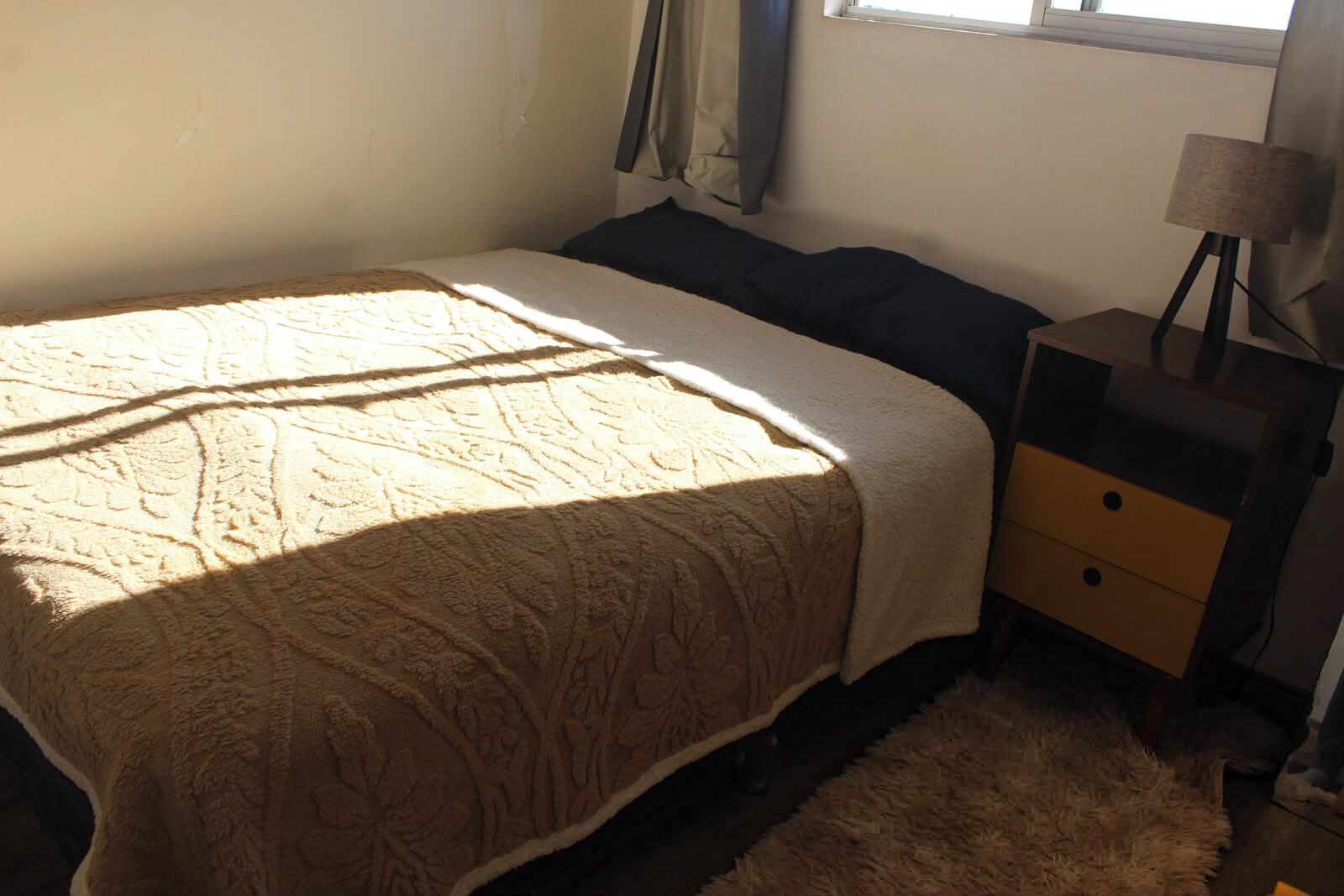Un dormitorio con cama, mesita de noche, lámpara y ventana con cortinas.