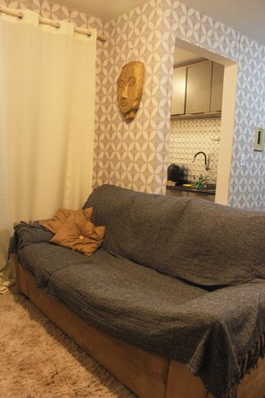 Una sala de estar con sofá, un papel tapiz estampado y una cocina visible a través de un umbral abierto.