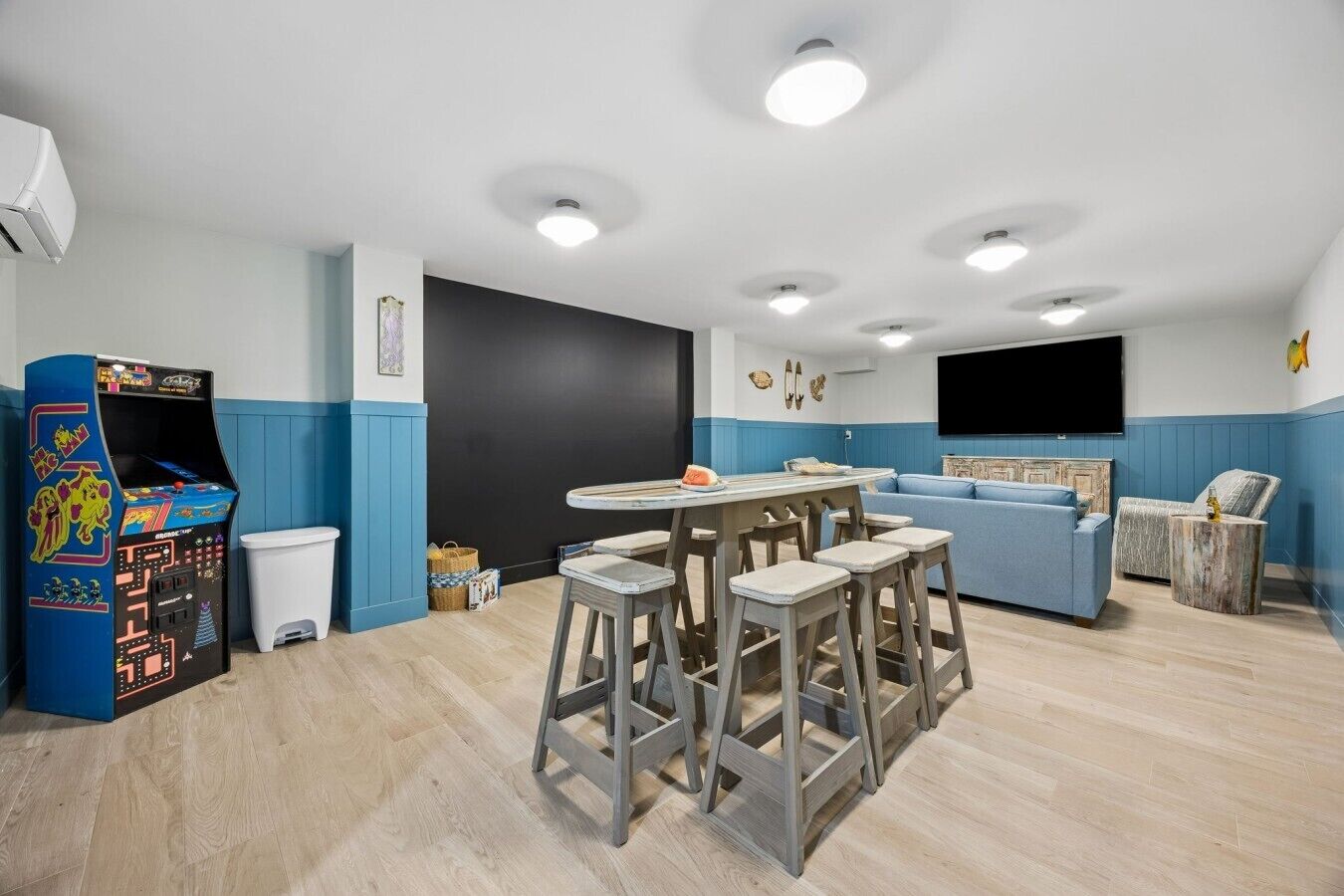 A room with a gaming console, a table with stools, a sofa, and a television.