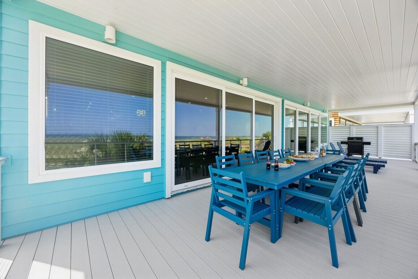A covered outdoor dining area with a long table and blue chairs, large windows, and a view of the ocean.