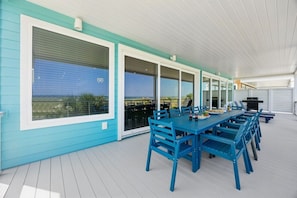 A covered outdoor dining area with a long table and blue chairs, large windows, and a view of the ocean.