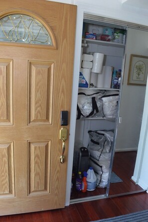 Una puerta de madera con un panel de vidrio y una cerradura electrónica. En el interior, hay un armario con estantes que contienen papel higiénico, productos de limpieza y bolsas.