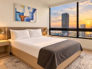 A hotel room with a large bed, a nightstand, a wall-mounted lamp, a painting, and a view of a city skyline at sunset. - Spacious 1BR Condo in Downtown Miami | Sleep 4  | Free Parking (Miami)