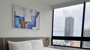 A modern bedroom with a large bed, a nightstand, a wall-mounted abstract painting, and a view of a cityscape through a large window. - Spacious 1BR Condo in Downtown Miami | Sleep 4 (Miami)