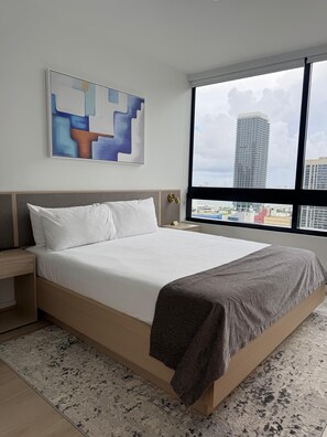 A modern bedroom with a large bed, a nightstand, a wall-mounted abstract painting, and a view of a cityscape through a large window. - Spacious 1BR Condo in Downtown Miami | Sleep 4 (Miami)