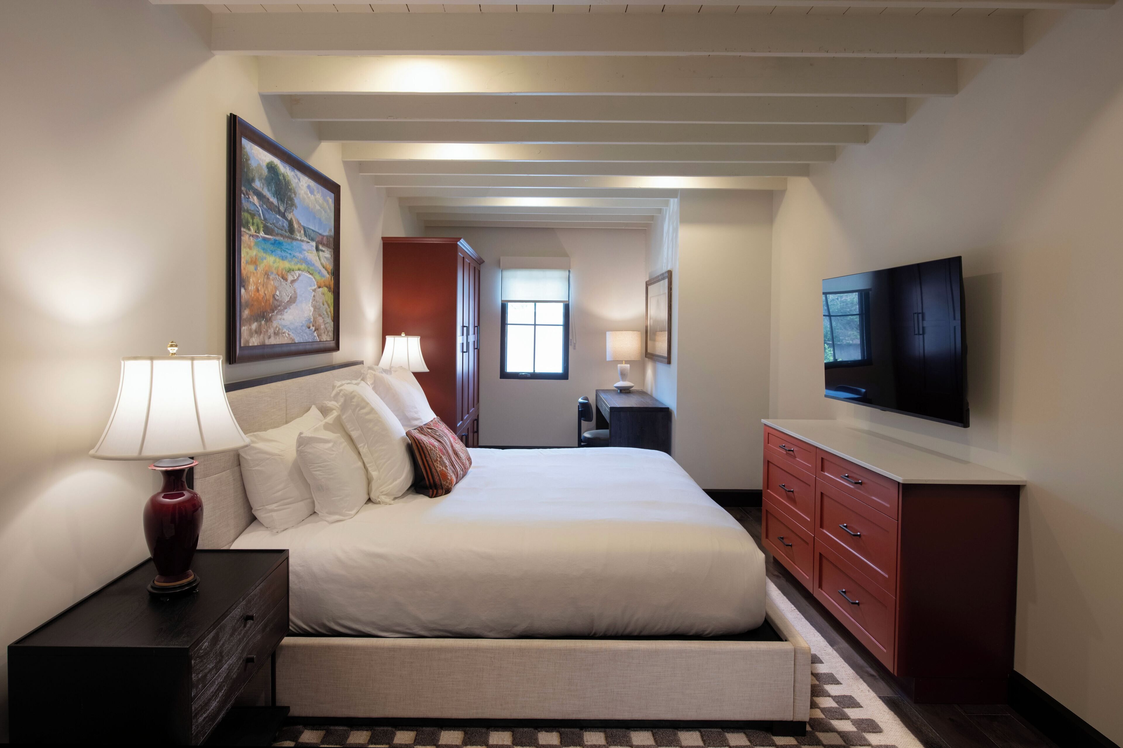 Quarto de hotel com cama, mesas de cabeceira, televisão, quadro e vista para o corredor.