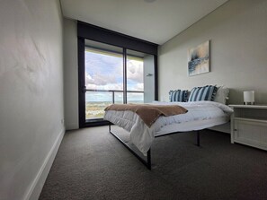 A bedroom with a bed, a nightstand, a window with a view, and a painting on the wall. - Sydney Olympic Park Panorama 3B2B by WinsonHome (Sydney Olympic Park)