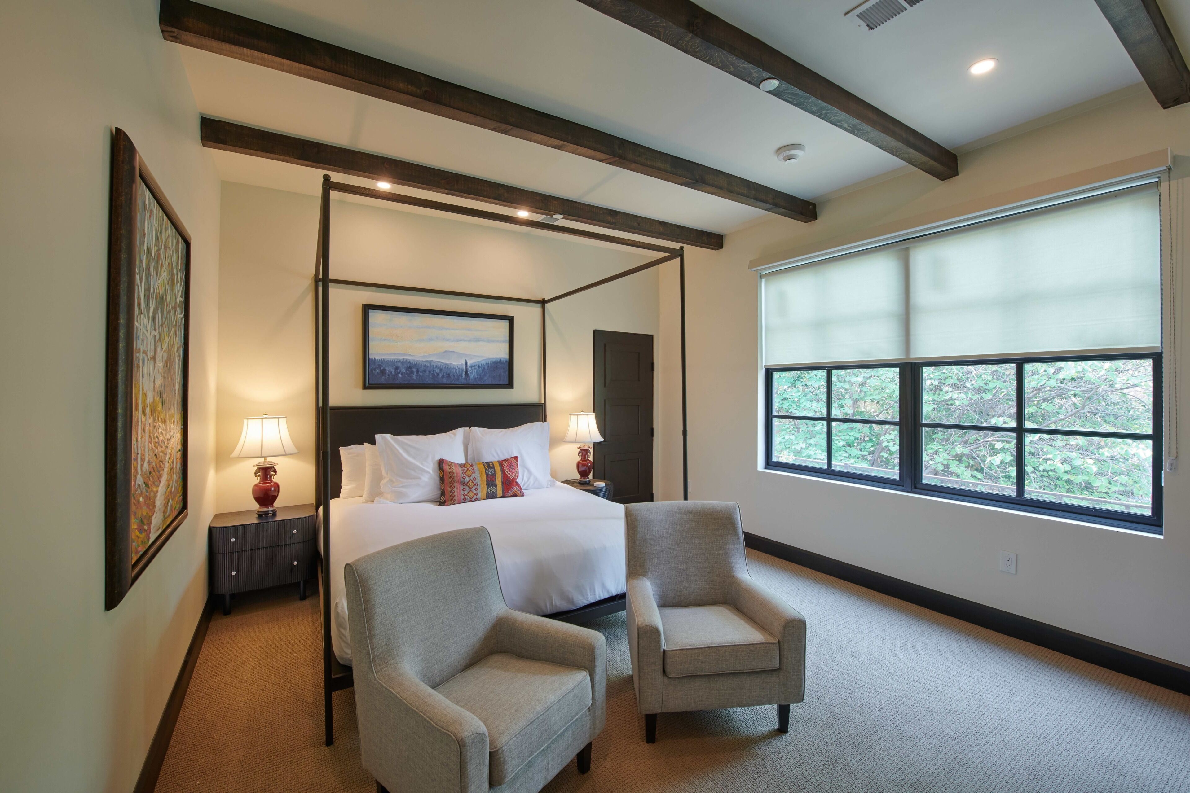 Quarto de hotel com cama de dossel, duas poltronas, uma janela ampla com vista e um quadro na parede.