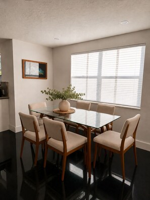 A dining area with a glass table and wooden chairs, a potted plant, and a window with blinds. - Newly refurbished, private home with open layout and backyard (Sarasota)