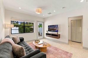 A modern living room with a sofa, coffee table, TV, and a view of greenery outside. - Renovated 1BR w Private Garden & Laundry (Miami Shores)