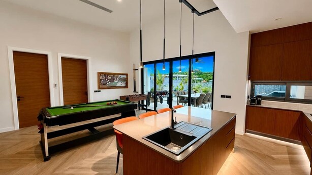 A modern kitchen with a central island, a pool table, and a dining area with a view. - Villa Amara - Viluma Villas (Hua Hin)