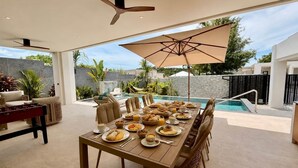 A dining area with a pool view, a table set for a meal, and a ceiling fan. - Villa Amara - Viluma Villas (Hua Hin)