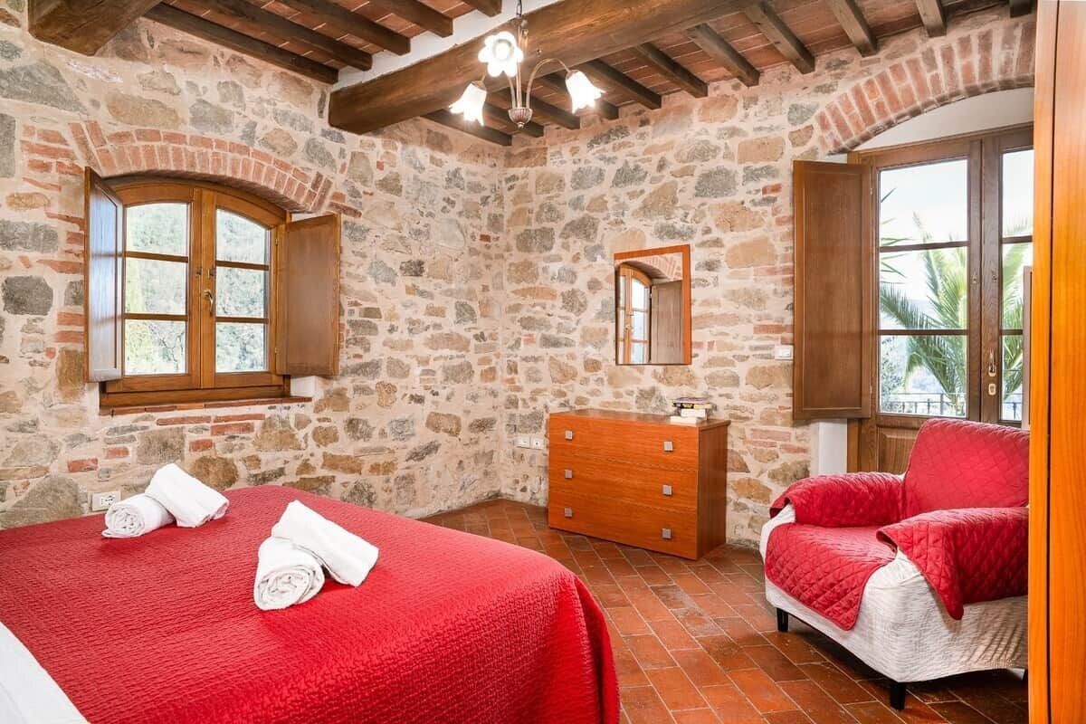 Une chambre aux murs de pierre, avec un lit, une commode et un fauteuil rouge.