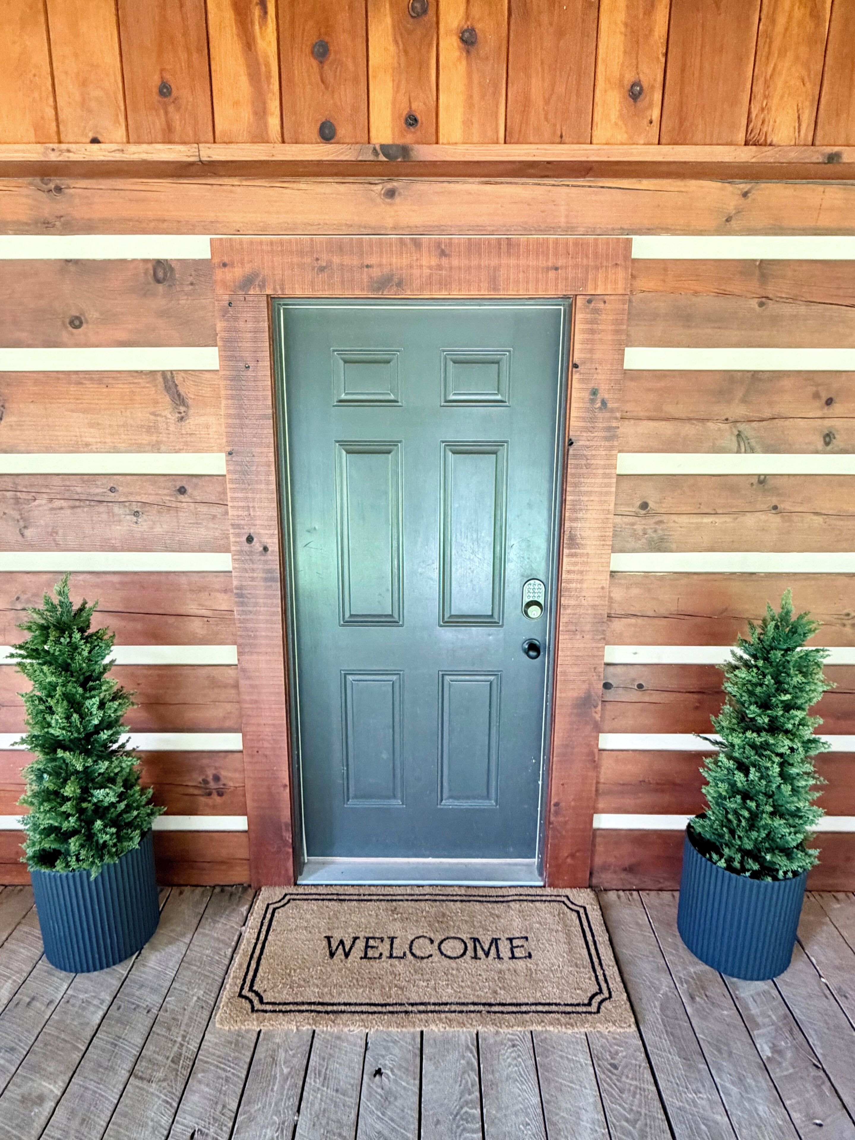 A wooden door with a welcome mat, two potted plants, and a wooden wall with horizontal planks.