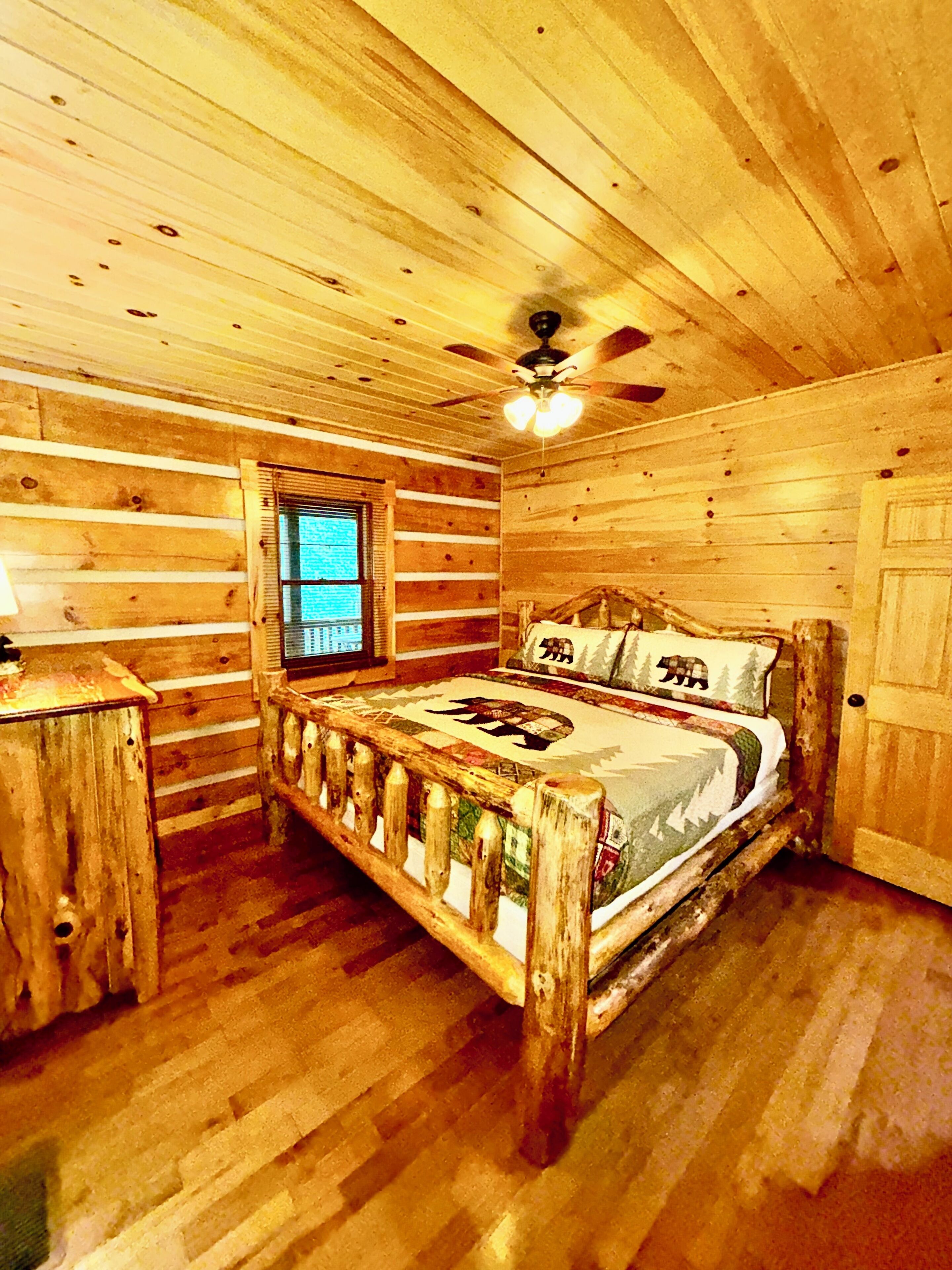 Ein Schlafzimmer einer Holzhütte mit einem Bett, einem Fenster, einer Tür und einem Deckenventilator.