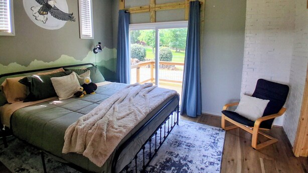 A bedroom with a bed, a chair, a wall mural, and a view through a window. - Sunrise over the lake and mountains...  (Albemarle)