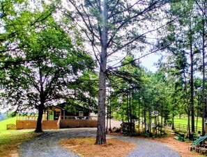 A rustic cabin with a wooden deck, surrounded by a forested area with a gravel path leading to it.