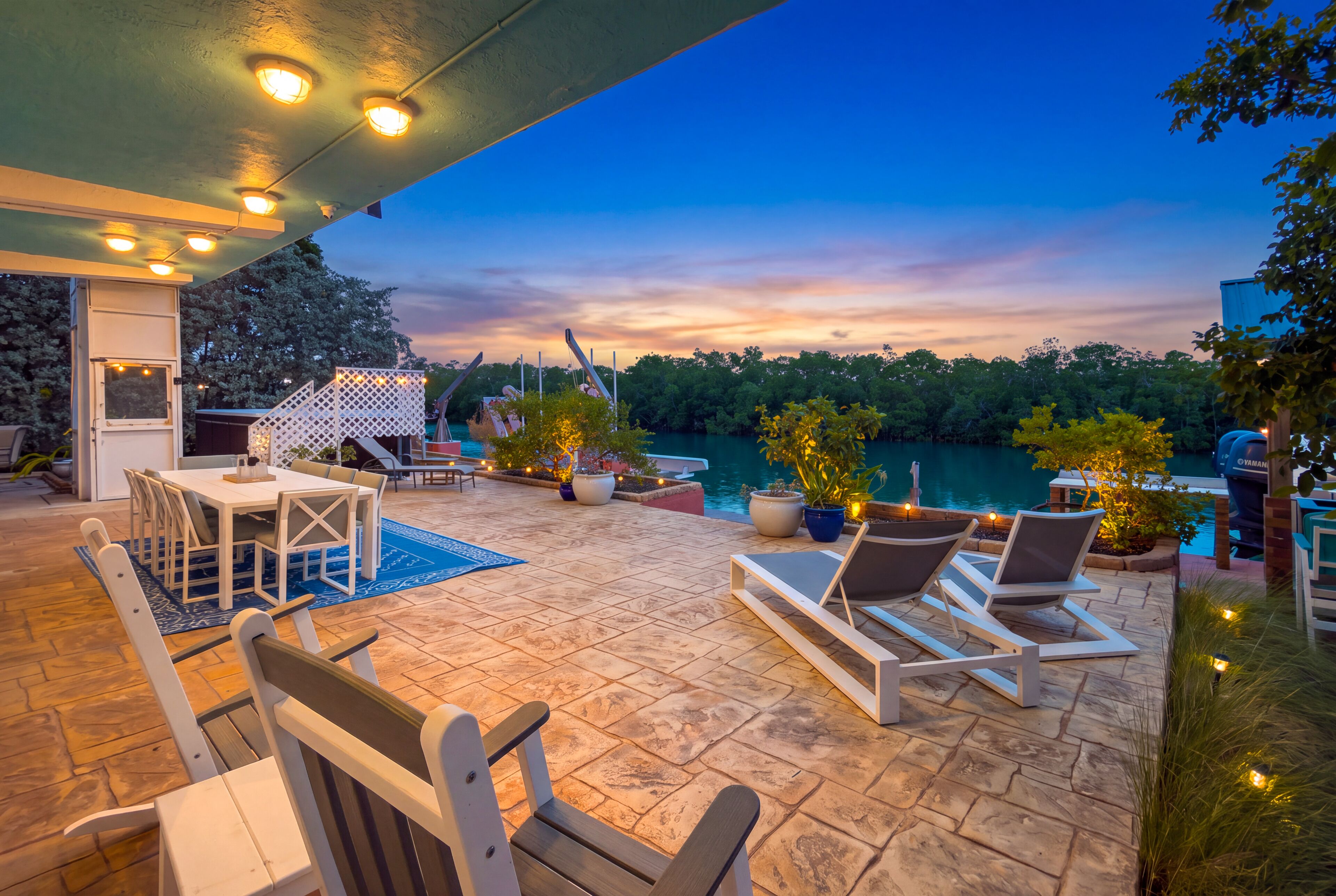 An outdoor patio with a dining area, lounge chairs, and a view of a body of water at dusk.