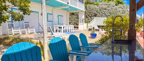 A poolside area with blue chairs and a table, a covered seating area, and a building with a balcony.