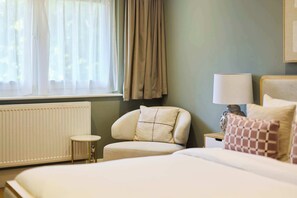 A hotel room with a bed, a chair, a small table, a lamp, and a window with curtains. - Well Placed in the Wood (Greater London)