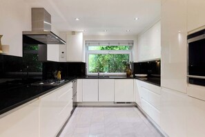 A modern kitchen with white cabinetry, black countertops, a stainless steel range hood, and a window with a view of greenery. - Well Placed in the Wood (Greater London)