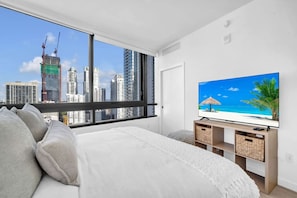 A modern bedroom with a large window, a bed, a flat-screen TV, and a view of a cityscape. - District 225 - Brand New Downtown Apartment (Miami)