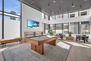 Un espace piscine intérieur moderne, doté d’une table de billard, de coins salon et de grandes fenêtres offrant une vue sur le paysage urbain.