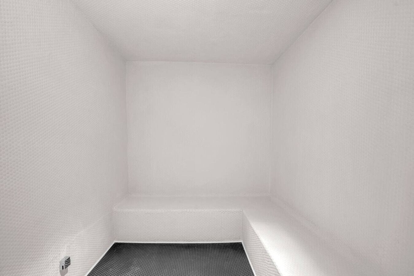 Ein weißer, leerer Raum mit strukturierter Wand, einem kleinen Fenster und einem Lüftungsgitter unten.