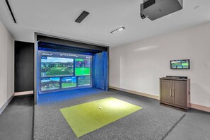A room with a large screen displaying a golf game interface, a small cabinet, and a carpeted floor. - District 225 - Brand New Downtown Apartment (Miami)