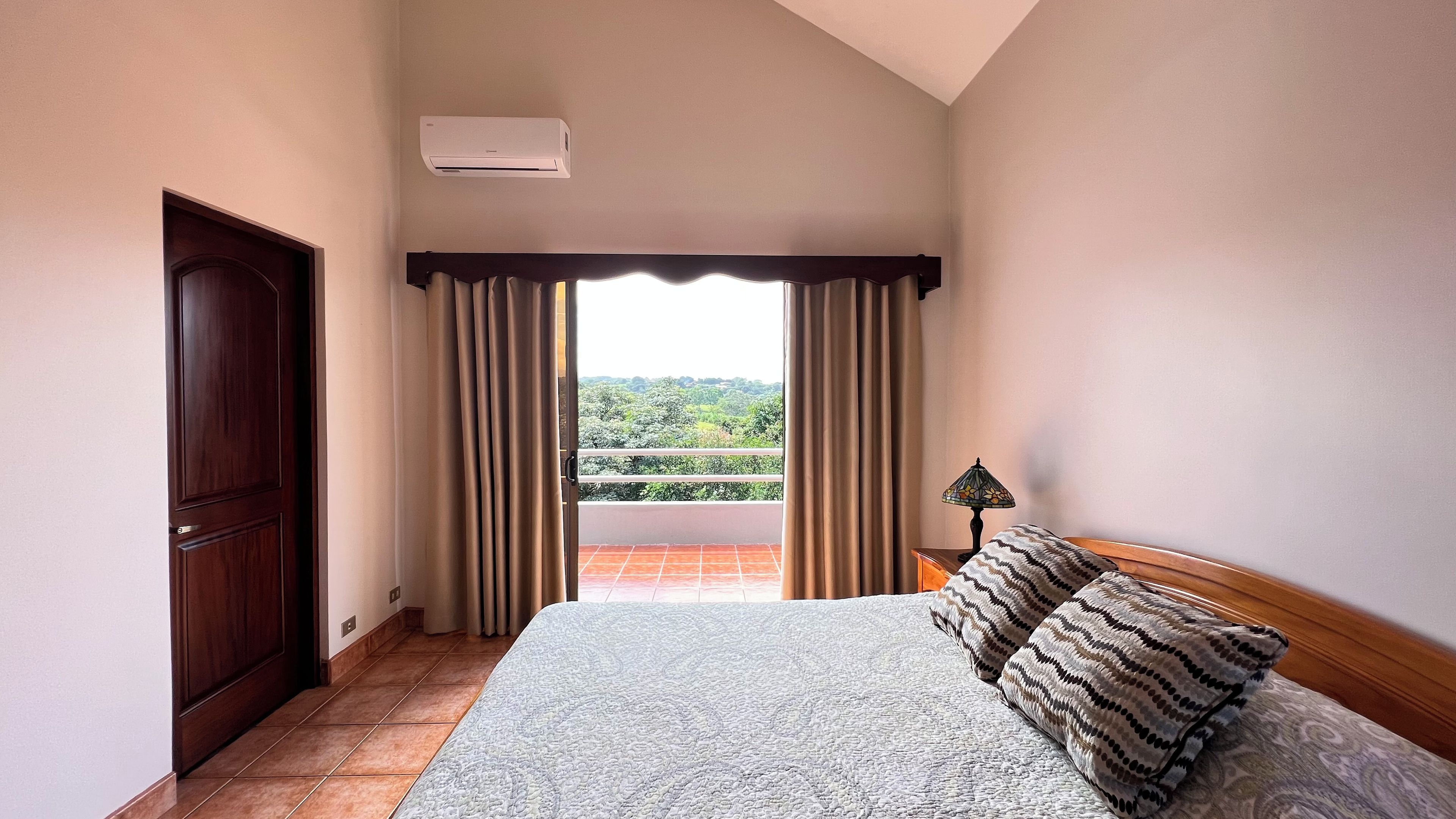 A bedroom with a large bed, a nightstand with a lamp, a window with curtains, and an air conditioning unit.