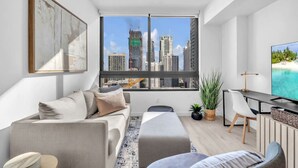 A modern living room with a large window, a sofa, a desk, and a television. - Brand New Building & Amazing Amenities (Miami)