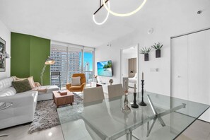A modern living room with a glass dining table, white sofas, a green accent wall, and a large window with a city view. - Basquiat Brickell Luxury Retreat (Miami)
