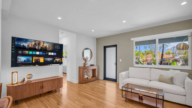 Uma sala de estar moderna com televisão de tela plana, piso de madeira, sofá branco e janela com vista para o verde.