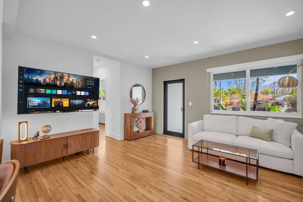 Ein modernes Wohnzimmer mit Flachbildfernseher, Holzboden, einem weißen Sofa und einem Fenster mit Blick auf Grünflächen.