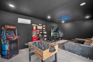 Um quarto com mesa de pebolim, jogos de fliperama, uma TV grande e um sofá.