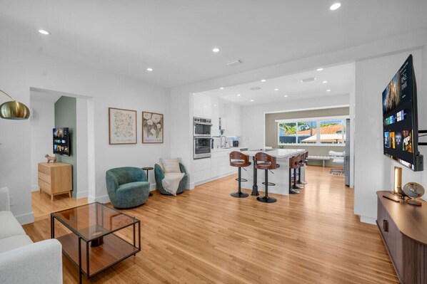 A modern living room with a glass coffee table, a teal armchair, and a kitchen area with a bar. - The Palm Cozy Lounge Pool, Private Theater & Games (Miami)