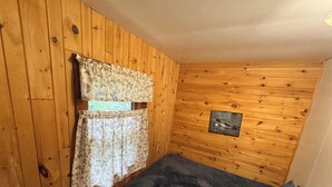 Una stanza piccola con pareti in legno, un letto con coperta scura, tende floreali e un quadro appeso al muro.