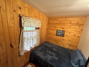 A small room with wooden walls, a bed with a dark cover, floral curtains, and a framed picture on the wall. - 2 Bedroom cabin in the heart of the Hiawatha National Forest... (Manistique)