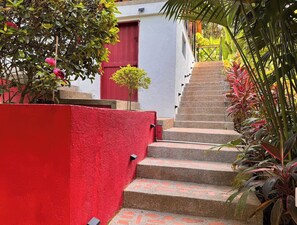 Eine Treppe führt zu einem Gebäude mit einer roten Tür, umgeben von Grünpflanzen und einer roten Mauer.