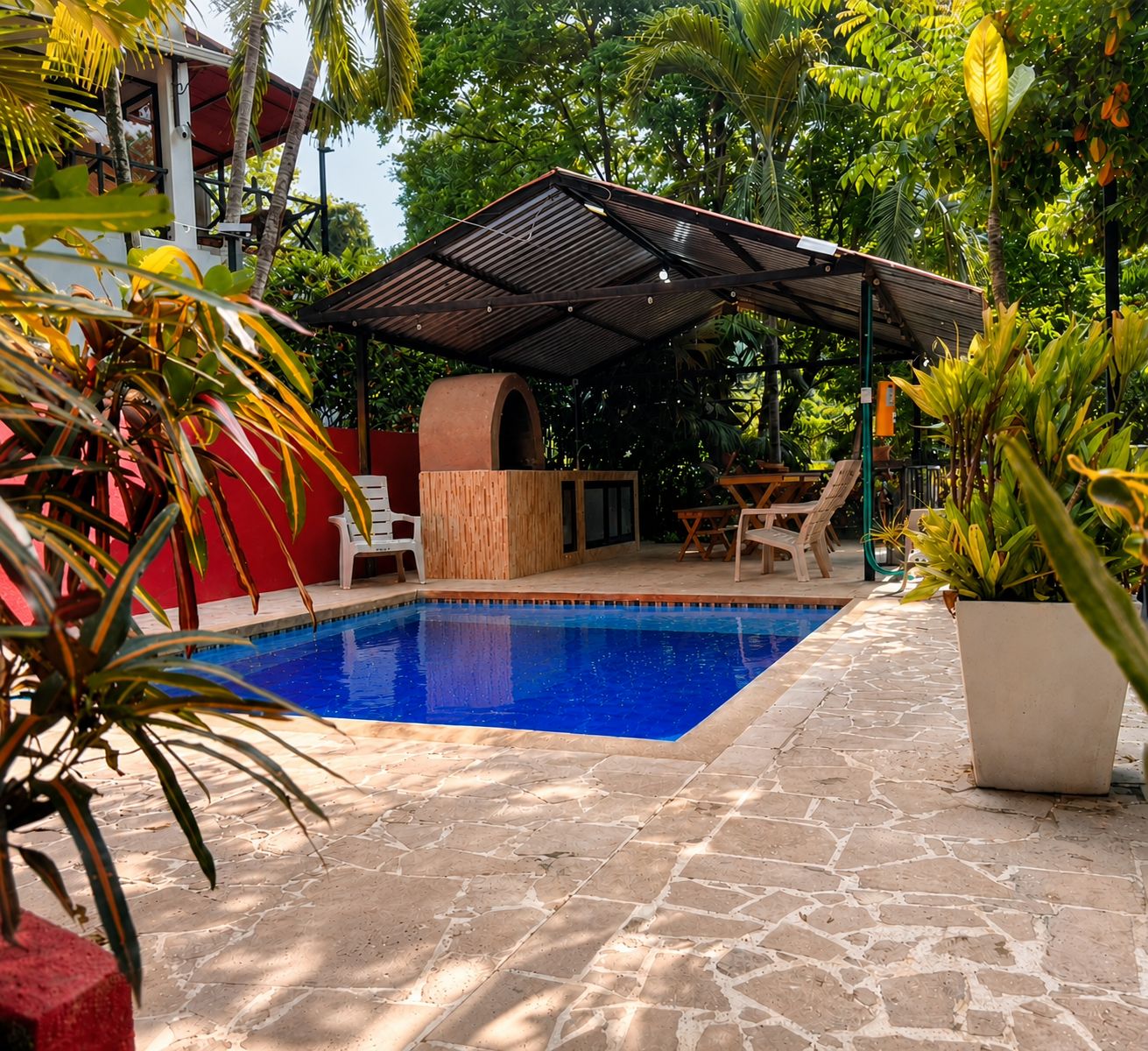 A swimming pool with a covered seating area, surrounded by stone flooring and lush greenery.
