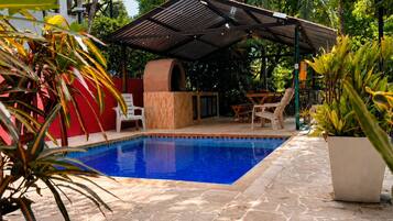 A swimming pool with a covered seating area, surrounded by stone flooring and lush greenery.