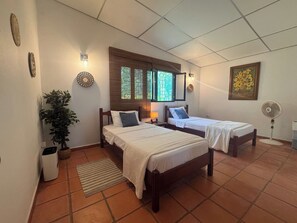 A room with two beds, a potted plant, a wall-mounted light fixture, and a window with blinds.