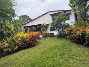 Una casa moderna con fachada blanca y techo inclinado, rodeada de un jardín cuidado con diversas plantas y un césped.