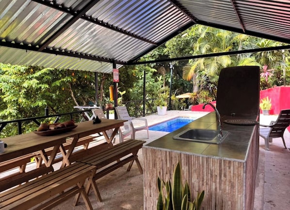 An outdoor covered area with a pool, wooden furniture, and a bar.