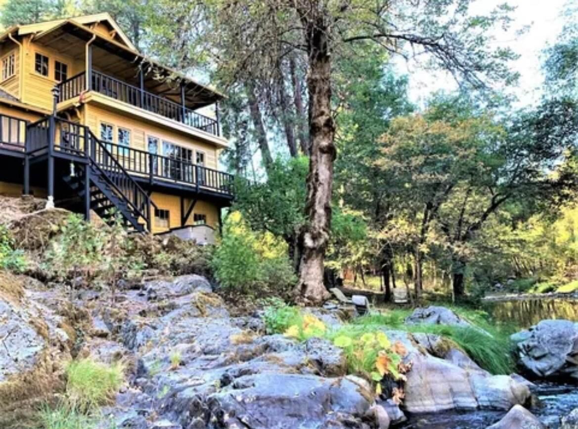 Una cabaña de madera de dos pisos con balcón, rodeada por un paisaje natural con árboles y un arroyo pedregoso.