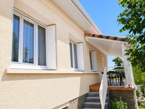Ein Gebäude mit einer beige gefärbten Fassade, weißen Fensterläden und einer Treppe zu einem überdachten Außenbereich mit einem Metallgeländer.