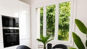Eine moderne Küche mit weißen Schränken, einem runden Esstisch, schwarzen Stühlen und einem großen Fenster mit Grünpflanzen draußen.