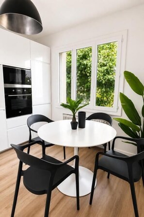 Eine moderne Küche mit weißen Schränken, einem runden Esstisch, schwarzen Stühlen und einem großen Fenster mit Grünpflanzen draußen.