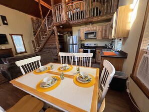 Un coin repas avec une table en bois dressée pour quatre personnes, entourée de chaises blanches. Un escalier avec des rampes en bois mène à un niveau supérieur.